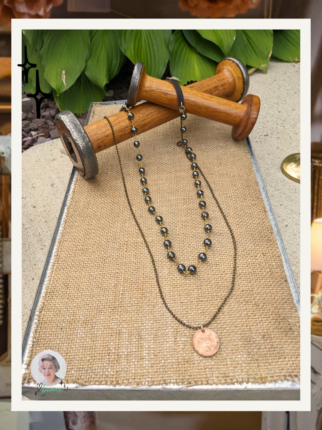 Layered necklaces with dark beaded chain and antique-style coin pendant, styled on burlap with wooden spools and greenery in the background.