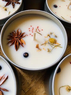 Small tins of creamy white candles topped with star anise, dried flowers, and spices, arranged closely together on a wooden surface.